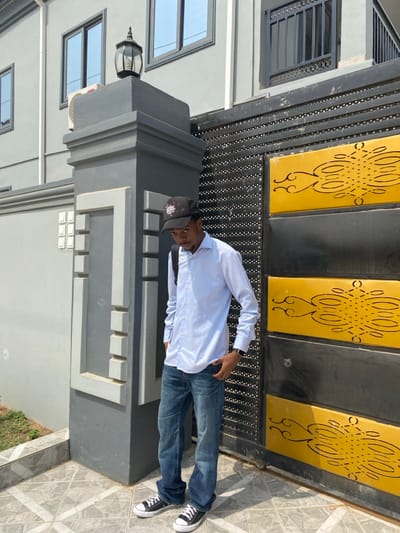 This image, taken in Joma, Ghana, portrays a young adult male standing outdoors in daylight. He is positioned slightly left of center, facing forward with his head tilted downwards and to his right. The individual wears a black baseball cap with a white and red logo, a light blue long-sleeved collared shirt, blue denim jeans, and black Converse-style sneakers with white laces. A black backpack strap is visible over his left shoulder, and a black wristwatch is present on his left wrist. Both of his hands are in the front pockets of his jeans.
To the left of the male is a dark grey concrete pillar, topped with a black, ornate outdoor lamp featuring a clear glass enclosure. The pillar's visible side is adorned with lighter grey rectangular recesses. To the male's right is a black and yellow metal gate, composed of horizontal black panels with rows of small circular studs and two wider yellow panels featuring elaborate black scrolling patterns.
Behind the pillar and the male stands a light grey multi-story building with several rectangular windows, framed in dark material, reflecting the sky. An outdoor air conditioning unit is mounted on the building wall, situated below one of the windows. The ground in the foreground consists of light grey square tiles laid in a diagonal pattern. A small patch of green grass is visible in the lower-left corner of the frame.