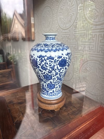 A close-up, eye-level shot captures a blue and white porcelain vase with intricate floral patterns, displayed on a polished wooden table in Xicheng District, China. The vase rests on a circular wooden stand, and its reflective surface mirrors the surrounding environment. The background features a textured wallpaper with a repeating circular motif and geometric patterns. To the left, a reflection in a glass surface reveals a glimpse of a traditional Chinese building with a gabled roof, surrounded by greenery. The scene is lit with ambient light, creating soft shadows and highlights on the objects and surfaces.