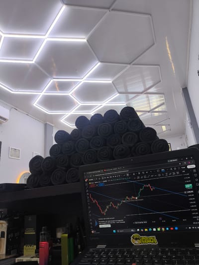 The image shows a desk setup with a laptop displaying a financial trading chart, stacks of rolled-up black towels, and various other objects on the desk and shelf. The ceiling features a modern hexagonal lighting design with integrated LED lights. In the background, there's a framed certificate on the wall and what appears to be an air conditioning unit. The laptop's screen shows a chart with Japanese candlesticks, indicating active trading. Below the towels, there are several vape devices and a box of "HAIR TREATMENT SHAMPOO." The overall environment suggests a personal workspace, possibly in a home or small business setting in Miruektaman, Indonesia. There are no people visible in the image.