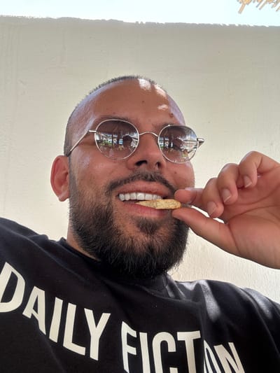 A man is shown from the chest up, looking directly at the camera. He is wearing a black t-shirt with white lettering that reads "DAILY FICTION". The man has a shaved head and a beard, and he is wearing round, silver-rimmed sunglasses. His right hand is raised, and he is holding a biscuit to his mouth, as if about to take a bite. The man's teeth are visible, and they appear to be white and even. The background is a plain white wall, and there is some natural light coming from the top left of the frame. The image appears to have been taken outdoors, possibly in a tropical setting, given the reflections in the man's sunglasses which show palm trees and umbrellas. However, there is no indication of Baliovtsi, Bulgaria in the image.