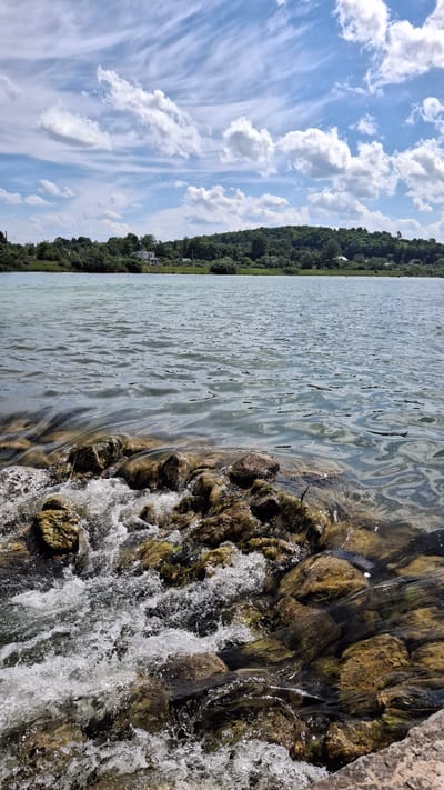 The image displays an outdoor landscape scene within the general vicinity of Ветеран Снт, Russia, under a bright, partly cloudy sky. The composition is horizontally layered, featuring turbulent water and rocks in the foreground, calmer water in the midground, a forested landmass with scattered buildings in the background, and a dynamic sky above.
In the immediate foreground, a section of water is vigorously agitated, forming white rapids as it flows over numerous partially submerged and exposed rocks. These rocks exhibit irregular shapes and patches of greenish-brown growth, indicative of algae or moss. The water in this area appears frothy and white due to the rapid movement.
Beyond the rapids, the main body of water extends across the midground. This section is calmer, displaying ripples and reflecting the sky, with a muted greenish-blue color.
The distant bank across the water, located towards the background, is densely covered with lush green trees and foliage, forming a continuous line of vegetation. Several light-colored structures, identifiable as houses or small buildings, are visible nestled within or peeking above the tree line on this distant bank. A faint linear feature, possibly a road or railway track, runs horizontally behind the initial tree line.
The sky occupies the upper portion of the image and is bright blue, adorned with a variety of clouds. Both wispy, high-altitude cirrus clouds and more distinct, fluffy cumulus clouds are present, scattered across the frame. No individuals or discernible human activity are depicted in the scene.