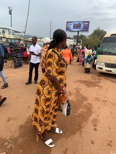A busy outdoor market scene in Nyanya, Nigeria, is depicted in this vertical, full-shot image. The ground is dusty and uneven, with patches of wetness suggesting recent rain. A light-colored bus is partially visible on the left, indicating it's a transportation hub or a stop along a road. Several people are present, engaged in various activities.
In the foreground, a woman wearing a vibrant yellow and orange patterned dress stands facing away from the camera. She has dark braided hair and is carrying a brown bag over her shoulder. White sandals are visible on her feet. Behind her, a man in a white short-sleeved shirt and dark pants stands looking at his phone.
The middle ground reveals more activity. A stall is set up with various items for sale, including bags and what appear to be sunglasses. Several people are gathered around, possibly shopping or waiting. One person in an orange shirt is also visible. Further back, a billboard with text is prominently displayed. The billboard features the word "Royal" in large blue lettering and below it, "ASCEND THE THRONE" in white. The overall atmosphere appears lively and bustling, typical of a market day. The sky is overcast, suggesting a cloudy day.