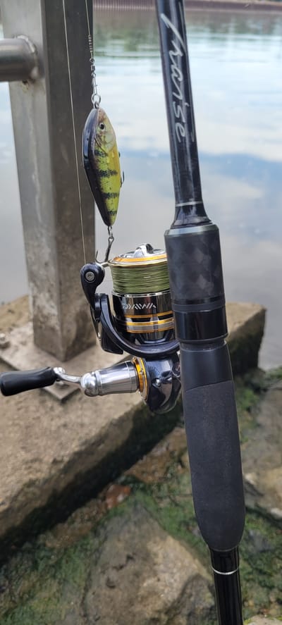 A fishing rod with a reel and lure hangs vertically in the foreground, positioned in front of a blurred background of water and some concrete structures, suggesting it is near a body of water. The fishing rod has black grip and silver accents, and the reel is black and gold with green fishing line. A lure, designed to resemble a yellow and black fish, is suspended from the fishing line above the reel. The scene appears to be outdoors, possibly at a waterfront park or pier in Klein-Krotzenburg, Germany, given the presence of water and man-made structures. The lighting is natural, suggesting it is daytime.