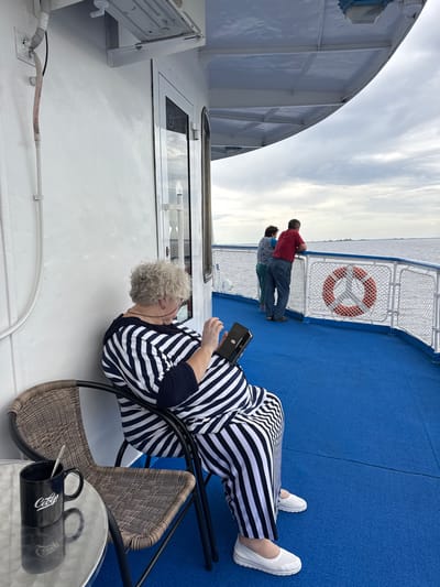 An elderly woman with curly gray hair is sitting on a wicker chair on the deck of a vessel. She is wearing a black and white striped jumpsuit and white slip-on shoes. She is looking down at a black electronic device in her hands. Next to her chair is a small round table with a black mug and a spoon. In the background, a couple is standing near the railing, looking out at the water. A life preserver is attached to the railing. The deck is painted blue. The sky is overcast with clouds. The location appears to be on a body of water, possibly near Krasny Bor, Russia, given the context provided.