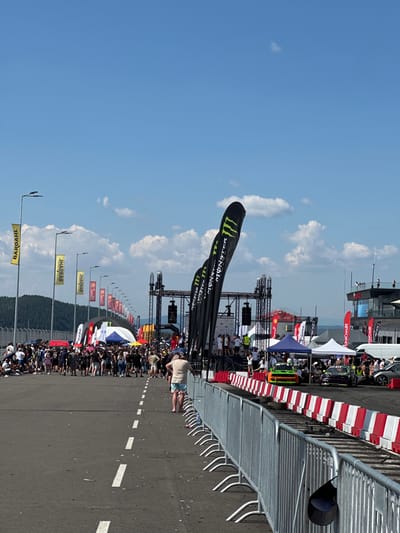 An outdoor event, likely a race or festival, is taking place on a bright, sunny day in Dospey, Bulgaria. The scene is crowded with spectators, many of whom are standing or walking along a paved area. Several large flags, some bearing the "Monster Energy" logo, are prominently displayed. A stage structure with lighting equipment is visible in the background, suggesting entertainment or presentations. Barriers made of metal fencing and red and white concrete blocks line the event area, separating the spectators from a track where race cars are present. One car, a green sports car, is parked on the track, while another, a darker colored race car, is also visible. Tents and booths are set up, indicating vendors or event support areas. The environment includes a clear blue sky with scattered clouds and a distant view of trees on a hillside.