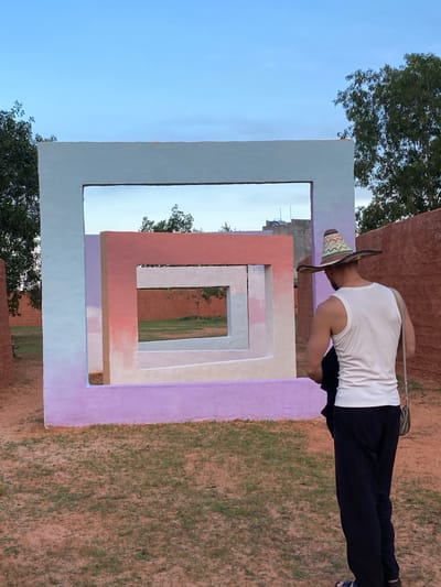 An adult male wearing a white tank top, dark pants, and a patterned sombrero stands facing away from the camera, looking at a colorful, abstract sculpture. The sculpture is composed of nested square frames painted in shades of light blue, pink, and purple. The background features sparse trees and a wall of reddish-brown material. The ground is covered in sandy soil with patches of green grass. The scene appears to be outdoors in a desert-like environment, potentially in Mui Ne, Vietnam, known for its sand dunes and artistic installations. The lighting suggests it is late afternoon or early evening.