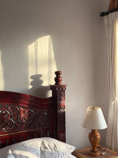 The image shows a bedroom interior in Abang, Indonesia, bathed in the warm glow of natural light. A dark red wooden bed frame with intricate carvings is the dominant feature, with a white pillow resting on it. To the right, a bedside table made of woven material holds a cream-colored lampshade on a wooden base, with its cord plugged into an unseen outlet. A sheer white curtain hangs to the right, partially drawn. A shadow cast by the curtains falls on the wall behind the bed, creating a dappled light effect. The overall scene depicts a serene and cozy bedroom environment.