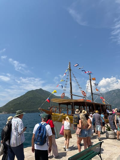 In Perast, Montenegro, a group of tourists is gathered on a paved walkway next to the Bay of Kotor, awaiting boarding of a yellow tour boat. The boat is equipped with two masts adorned with numerous colorful flags and nautical signals. The vessel, with its wooden deck and covered seating area, is positioned to embark passengers. A lush, green, tree-covered mountain rises from the water in the background, under a bright blue sky dotted with white clouds. Several individuals are visible, some carrying backpacks and wearing casual summer attire, indicating they are likely on vacation. The overall scene depicts a sunny day at a popular tourist destination.