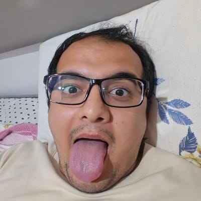 A close-up, forward-facing selfie of a person lying on their back on a bed. The person is wearing black-rimmed glasses and has their mouth open with their tongue extended. The background includes a pillow with a blue floral pattern, a white wall, and a glimpse of a striped pillow and a pink and white striped blanket. There are no discernible landmarks to indicate the specific location within Küçükçekmece, Turkey.
