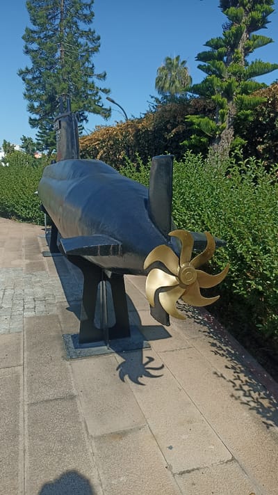 A black, model submarine is displayed outdoors on a paved walkway in Yenişehir, Turkey, under a clear blue sky. The submarine is elevated on black metal stands and has a prominent brass propeller at its stern. Green foliage, including trees and bushes, surrounds the display. Shadows are cast on the ground from the submarine and surrounding vegetation, indicating a sunny day. The scene appears to be a public park or memorial site.