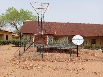 The image shows a satellite dish, a tall metal tower with a square antenna, and a set of solar panels on the ground. The solar panels are enclosed by a wire fence. In the background, there is a building with a rusty corrugated metal roof. The building has windows with metal bars and a door. The image appears to be taken in the courtyard of a building. There are dry leaves and dirt on the ground. A metal gate is lying on the ground in the foreground. The scene is located in Rumde Waziri, Nigeria, at the coordinates 9.184 latitude and 12.521 longitude.