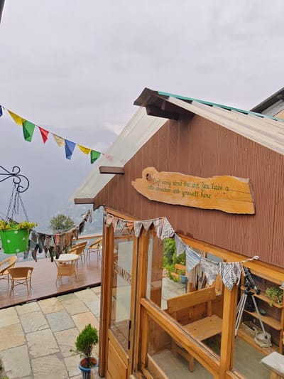 The image depicts an outdoor seating area at a cafe or guesthouse in Prini, India, with a mountainous backdrop. In the foreground, a wooden deck is laid with stone tiles. On the deck, several wicker chairs and a circular table are arranged. A green planter with a plant hangs from a black metal hook. Prayer flags in various colors are strung across the upper portion of the image, with another string of fabric bunting hanging lower down.
The main structure is a wooden building with a corrugated brown exterior and a translucent roof. A wooden sign with a carved quote, "Don't worry about the wifi. You have a better connection with yourself here," is affixed to the building's wall. A glass-fronted wooden structure, possibly an extension or seating area, is visible to the right, with a wooden bench inside. A telescope on a tripod stands in this area.
The overall environment suggests a tranquil, possibly slightly overcast day, with lush greenery and distant hills visible. The scene is tidy and well-maintained, with decorative elements contributing to a bohemian or spiritual atmosphere.