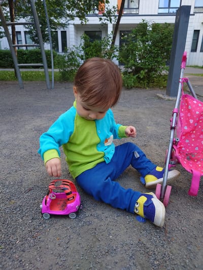 In an outdoor urban environment, likely a playground in Kommunarka, Russia, a young child is seated on a gravel surface. The child is wearing a long-sleeved shirt, green and blue, and blue pants. On their feet are blue and yellow sneakers with character embellishments. To the child's left is a small pink toy car. To the child's right is the frame of a pink doll stroller. In the background, a modern building with white and orange brickwork is visible, along with green foliage and parts of a playground structure. The overall scene depicts a moment of childhood play.