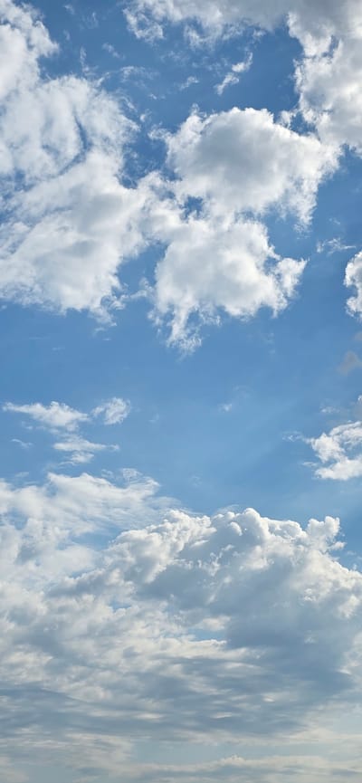This image depicts a bright, partly cloudy sky with variations in cloud density and altitude. The sky is a clear, vibrant blue, indicating daylight conditions. Puffy, white cumulus clouds are scattered throughout the upper portion of the frame, some appearing wispy and others more substantial. Towards the bottom of the image, a thicker layer of stratus clouds is visible, appearing lower in the sky and exhibiting more texture and shadow. Sunlight is filtering through the clouds, creating areas of brighter illumination and softer shadows. The overall impression is that of a dynamic sky on a pleasant day. There are no people, subjects, objects, or actions visible in the scene, as it is solely a sky photograph. The location is specified as Recaș, Romania, suggesting this is a view of the sky from that geographical area.