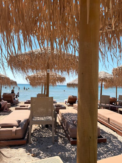 This media file captures a sun-drenched beach scene, likely in Greece, given the location context. The foreground is dominated by a rustic straw umbrella, its dried fronds creating a textured canopy against a clear blue sky. A thick wooden beam, part of the umbrella's structure, runs horizontally across the image, partially obscuring the view.
Beyond the umbrella, rows of lounge chairs and umbrellas line a sandy and pebbly beach. The chairs are predominantly a muted beige, some with cushions, arranged in neat rows. The sea is a tranquil blue, meeting the shore with gentle waves. In the water, several people can be seen wading or standing, enjoying the ocean.
The overall atmosphere is one of relaxation and leisure, indicative of a pleasant day at the beach. The bright sunlight and clear sky suggest fair weather, likely during the daytime, perhaps late morning or early afternoon, given the intensity of the shadows cast by the umbrellas and chairs on the ground. The scene evokes a sense of a typical summer vacation or a peaceful day by the sea. A small, faded blue sign with the number "3" is visible on one of the umbrella poles.