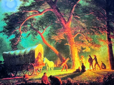 This image depicts a historical diorama or painting set in a wooded encampment scene, located in the city of Reus, Spain. The scene is illuminated by a prominent central campfire and ambient light suggesting a nighttime or twilight setting. On the left side, a large, canvas-covered wagon, clearly inscribed with "FOR OREGON" on its side, is parked on a dirt path. A light-colored horse stands beside the wagon.

In the foreground, two figures are seated on the ground, one dressed in red, the other a darker silhouette. Around the brightly burning campfire, several individuals are gathered. One figure is seen carrying a bundle of firewood towards the fire, contributing to its intensity. Other figures stand or sit, some silhouetted against the light, while others are illuminated with warm orange and yellow tones. Large trees with distinctive reddish-orange trunks and branches, strongly lit by the campfire, dominate the right and central portions of the scene, contrasting with the cooler blue and green hues of the less illuminated foliage and a patch of sky visible on the left side of the frame. The ground is a mix of dirt and sparse vegetation, with numerous small, bright flecks suggesting reflections or scattered light.