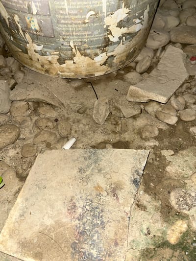 This image depicts a close-up view of debris and a damaged cylindrical object on the ground. The scene appears to be outdoors, with a rough, uneven surface composed of dirt, small stones, and broken concrete or plaster. The cylindrical object, possibly a container like a paint can or barrel, is covered in peeling paper labels with visible text. The labels are torn and weathered, suggesting exposure to the elements and age.

Visible text on the labels provides some context, including instructions and product information in what appears to be Indonesian. Phrases like "Cat telah siap langsung diguna" (Paint is ready to use), "Daya Sebar" (Coverage), "Waktu Kering sentuh" (Touch dry time), "Penyimpanan" (Storage), and warnings to keep away from children are legible. The location context provided is Kota Kuala Simpang, Indonesia.

The overall impression is one of neglect and disarray, with discarded materials and a generally unkempt environment. There are no people visible in the image, and the lighting suggests it might be daytime, though the quality of the light is difficult to determine precisely due to the close-up framing and the nature of the debris. The scene does not convey a specific event, but rather a static, abandoned-like setting. A white cigarette butt is also visible among the debris, adding to the sense of litter.