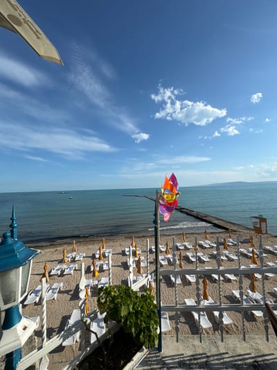 The image shows a beach scene in Balchik, Bulgaria. In the foreground, there is a white railing with decorative elements and a blue and white lamppost on the left. To the right of the lamppost is a planter with a green leafy plant. Behind the railing, rows of empty white lounge chairs and yellow umbrellas are arranged on a sandy beach. The beach stretches towards a calm, blue sea. A concrete pier extends out into the water from the middle of the frame. In the distance, a small white boat is visible on the sea. A colorful pinwheel is positioned on a pole on the beach, to the right of the center of the frame. The sky is a clear blue with some wispy white clouds. The overall impression is a serene and organized beach ready for visitors.