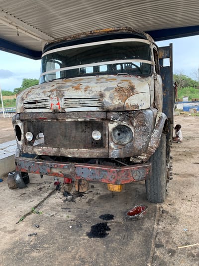 The image shows the front of a heavily rusted and damaged truck, possibly undergoing repair, under the partial shelter of a roof structure, likely a garage or workshop. The truck's front end is heavily corroded, with missing headlight lenses and deteriorated body panels. The grill is intact. Various parts and tools are scattered on the oily concrete floor around the truck. A child is atop the truck, appearing to interact with it, while the background reveals an outdoor environment. Based on the context, the image is likely taken in Gbeleko, Nigeria.