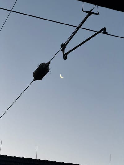 The image shows a clear sky with a thin crescent moon visible. Overhead power lines for a tram or train are silhouetted against the sky, with a pantograph arm extended. A large, dark insulator is attached to one of the power lines. In the foreground, the dark silhouette of a roof with several thin, vertical spikes is visible. The overall scene appears to be an urban environment, likely near tram or train tracks, during twilight or early evening. There are no people or animals present in the image. The location is identified as Witten, Germany.