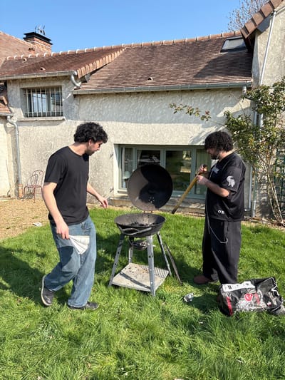 The scene is set in a sunny backyard of a residence in Bû, France, featuring green grass and a two-story house in the background with a light-colored, textured exterior and a terracotta-tiled roof.
Two males are present. The individual on the left has dark curly hair and is wearing a black t-shirt, blue jeans, and dark shoes. He holds a folded piece of white paper or newspaper in his right hand and is positioned near a charcoal barbecue grill, looking down towards it.
The individual on the right has dark curly hair and a beard, wearing a black graphic t-shirt, black baggy pants, and brown slippers. White wired earphones are visible around his neck and ears. He is actively engaging with the black kettle-style charcoal grill, holding a long wooden-handled tool into its interior, which contains charcoal remnants or unlit charcoal. The grill's lid is leaning against its base.
To the right of the grill, an open black bag labeled "BARBECUE" lies on the grass, alongside a small crumpled silver object. A light-colored metal garden chair is partially visible near the house wall on the far left.