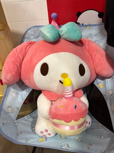 The image shows a close-up shot of two plush toys inside a light blue, patterned child's chair. The larger toy is predominantly pink with a white face and brown eyes, and a yellow object seemingly protruding from its mouth. The second, smaller toy is shaped like a cupcake, with pink and yellow layers, and a pink and white striped "candle." The chair has a light blue background with cartoon images of various animals and objects such as rabbits, cats, ice cream cones, and bananas. The mesh lining of the chair is visible beneath the toys. A red object with a white panda face is partially visible on the left side. The image was taken indoors, possibly in a retail setting. There is no indication of the specific location within Orlando, United States. No people are visible.
