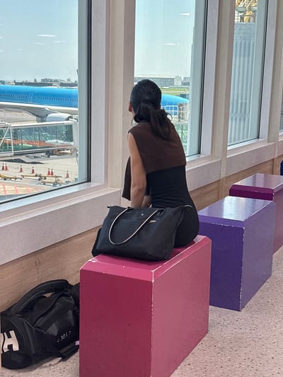 A woman with dark hair pulled back in a ponytail sits on a pink cube seat at an airport gate, looking out a large window at an airplane. She is wearing a dark sleeveless top with a brown scarf draped over her shoulders. A black handbag rests on her lap, and a black duffel bag is on the floor next to her. The airport gate features purple and pink cube seating arranged in a row. Outside the window, an airplane is visible at a gate, connected to the terminal by a jet bridge. The floor of the gate area is a terrazzo pattern. The scene suggests the woman is waiting for a flight at Jeju International Airport.