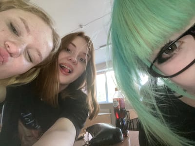 Three young women are captured in a selfie. The woman on the left has fair skin and freckles, with blonde hair. She is leaning into the frame, with her lips slightly parted. The woman in the middle has fair skin and brown hair, and she is smiling with her mouth open, her tongue visible. She is wearing a dark t-shirt with a graphic print. The woman on the right has light green hair and is wearing glasses. Her face is partially obscured by her hair, but her eye is visible, and she appears to be wearing dark eyeliner. She is wearing a dark top.

On a table in the foreground, there is a dark, textured handbag. Behind the women, a bottle of Coca-Cola is visible, along with what appears to be school desks and chairs. A window in the background lets in natural light. The overall environment suggests an indoor setting, possibly a classroom or a common area. There is no indication of the specific location in the city of Kaliningrad, Russia.