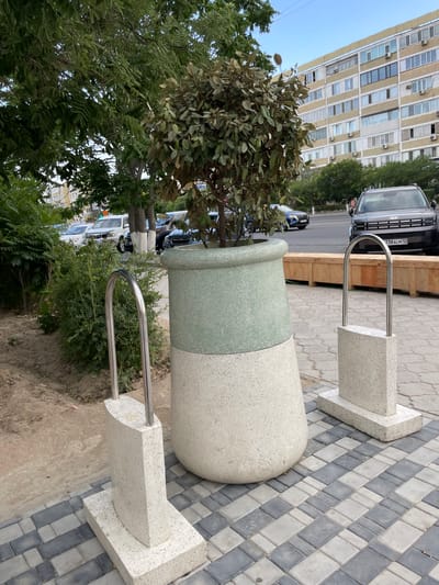 In the city of Aktau, Kazakhstan, a large, cylindrical concrete planter with a green top section stands on a patterned pavement. Two U-shaped metal bike racks, each with a concrete base, are positioned in front of the planter. Beyond the planter, a wooden bench is visible, and behind that, a dark-colored SUV with a license plate reading "738ALM 12" is parked. In the background, a multi-story apartment building with numerous windows and air conditioning units lines the street, with trees and more vehicles along the roadside. The scene appears to be an outdoor public space, possibly a pedestrian area or a streetscape.