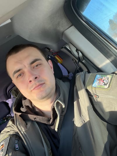 A male individual is positioned in the driver's seat of a vehicle, in what is identified as the city of Rogachovo, Russia. He has short, dark hair, brown eyes, and light stubble. He wears a black hooded garment beneath an olive-drab, tactical-style jacket with a dark, indistinct patch on the left arm. A black seatbelt is fastened diagonally across his chest. On the adjacent front passenger seat, an olive-drab tactical-style jacket or vest is draped, secured by the passenger-side seatbelt. This second garment features a rectangular patch on the right chest area depicting an off-road vehicle against a mountainous backdrop. The vehicle's interior presents a light grey headliner and pillar trim. Bright sunlight enters through the passenger window, revealing a clear blue sky and distant, bare trees outside, and creating prominent shadows within the cabin. Minor accessories are visible hanging behind the driver's headrest.