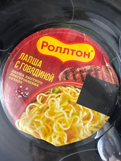 The image shows the lid of a cup of instant noodles with beef flavor from the brand "ROLLTON". The lid is red and yellow, with Russian text that translates to "Noodles with beef". There is a picture of a beef patty on the lid, and the noodles are visible at the bottom of the cup. The product is in a black plastic container, and the image appears to have been taken indoors. There is no visible information about the location being Novoaltaysk, Russia.