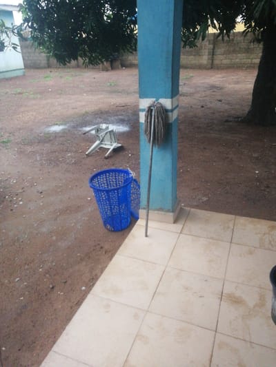 The image depicts an outdoor scene on a cloudy day, likely in Omupo, Nigeria. In the foreground, a tiled patio area is visible, with some dirt and debris scattered on the tiles. To the left of the patio, a blue plastic laundry basket with a second basket nearby sits on a dirt ground. Further back, a white plastic chair is overturned on the dirt. A blue painted pillar with white stripes stands on the patio, with a mop leaning against it. Behind the pillar, a large tree with dark green leaves partially obscures the view of a brick wall and a building. The ground is a mixture of reddish-brown dirt and patches of dry grass.