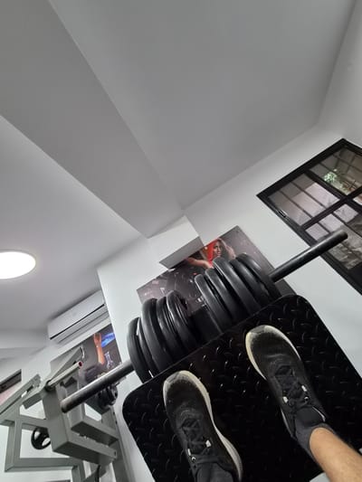 This image captures a dynamic, low-angle perspective of a gym or fitness room in Elena, Bulgaria. The main subject is a person's legs and feet, clad in black athletic shoes with white soles, positioned on the black textured footplate of a leg press machine. The machine's bar is heavily loaded with numerous black weight plates, indicating an active workout in progress. The equipment's frame is constructed from gray metal.

In the upper right corner, a large black-framed window with multiple panes offers a glimpse of the outside, revealing greenery and suggesting daylight. The room features stark white walls and ceiling, which, due to the skewed photographic angle, create strong geometric lines and shapes. On the wall behind the leg press, two posters are visible: one depicts a person, possibly a woman, wearing a red hooded top, while another shows a person with an arm raised.

Towards the lower right, a white wall-mounted air conditioning unit is installed, with the brand name "AUX" clearly visible on its front. A circular, recessed light fixture illuminates a section of the white wall in the lower-left area. The overall setting is brightly lit, consistent with a gym environment during the daytime. No specific emotions are conveyed, as the focus is primarily on the equipment and setting.