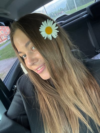 In the city of Chernushka, Russia, a young woman with long, blonde-brown hair is captured in a selfie taken from the backseat of a car. A single daisy is tucked behind her ear. The woman is wearing a black jacket, and her face is turned towards the camera, with a wide smile revealing her teeth. The car's interior is visible, including a dark gray seat. Outside the car, a building with red brick and multiple windows can be seen, along with a grassy area and a road. The background suggests an overcast day.