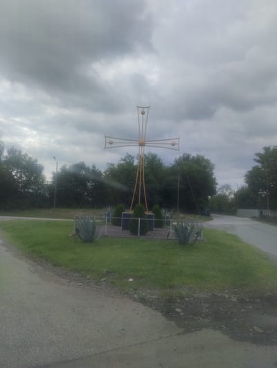 A large, golden-colored cross monument stands prominently in a grassy area. The cross is ornate, with spherical embellishments at its tips and center, supported by thin, angled legs. Surrounding the base of the cross is a low fence, and within this enclosure are several small, dark green conical plants and what appear to be large, spiky succulents. A paved road curves from the lower left of the frame towards the right, bordering the grassy area. In the background, a dense line of green trees forms a backdrop under a heavily overcast, cloudy sky. A single street lamp is visible to the left of the cross. The scene is static, with no people or animals present. The overall atmosphere suggests an outdoor location, possibly a roadside monument, in the region of Partskhanakanevi, Georgia.