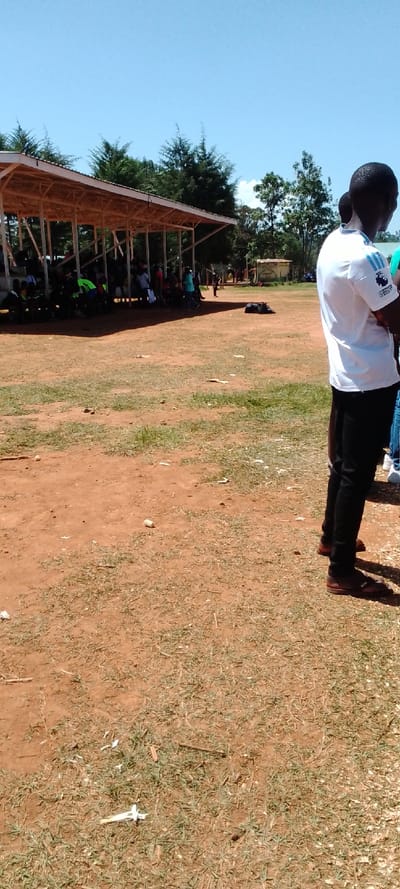 The image depicts a scene taken in Kipkarren, Kenya. It shows a group of people standing in a field under a large, wooden, covered structure. Several people are sitting on the ground under the structure, and some are visible walking in the distance. One man is standing in the foreground, facing away from the camera. He is wearing a white shirt with a blue design on the chest and black pants. The ground is a reddish brown dirt with patches of sparse green grass. There is a black bag lying on the ground in the middle of the field. The sky is clear and blue.