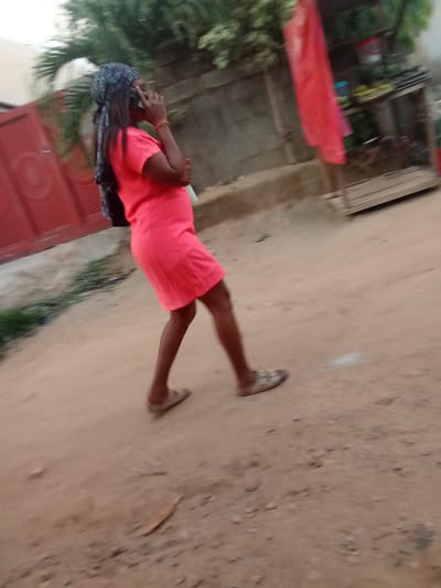 A medium-shot, motion-blurred photograph captures a woman walking on a dirt road in Kagini, Nigeria. The woman is wearing a vibrant pink dress and a dark headscarf with a patterned design. She is holding a mobile phone to her right ear with her right hand and her left arm is bent across her body. Her legs are visible from the knee down, with her left leg slightly in front of her right. She is wearing open-toed sandals with light-colored straps. The road is made of reddish-brown dirt and appears uneven. To the left of the woman, a section of a red wall or gate with a textured design is visible. In the background, blurred foliage suggests trees and greenery. To the right, a stall or shelf structure with items on it is partially visible. The overall impression is that of a candid shot of everyday life on a street in Kagini.