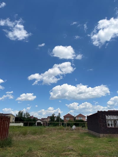 The image shows a residential area under a bright blue sky with scattered white cumulus clouds. On the right side of the frame, there is a grassy field with some dry, tall grass. Beyond the field, a row of houses can be seen, some of which appear to be under construction, with exposed brickwork and unfinished roofs. Trees and fences are interspersed between the houses. The sky occupies the left side of the image, filled with fluffy white clouds that cast soft shadows. The lighting suggests it is daytime, likely late morning or early afternoon, with clear weather. There is no visible text in the image. The scene depicts a suburban or semi-rural setting in Taganrog City District, Russia, with ongoing development or renovation of residential properties. The overall impression is one of a typical summer day.
