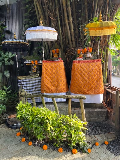 The image portrays an outdoor scene, likely in Ubud, Indonesia, with traditional Balinese Hindu offerings and decorations. Two shrines or altars are draped with patterned textiles—one in orange with a red border and another in a black and white checkered pattern. Flowers are arranged as offerings.
Bamboo stalks serve as a backdrop, interspersed with ceremonial umbrellas in yellow, white, and black and white. A wooden railing borders the scene, which is adorned with green foliage and scattered orange blossoms on a stone path.
