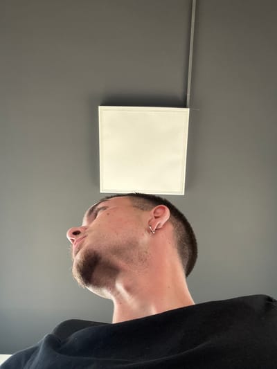 A low-angle shot shows the side profile of a young Caucasian male with a short buzz cut and a small silver hoop earring in his left ear. He is looking upwards. A plain white, rectangular light fixture is mounted on a dark grey ceiling above him. A thin silver cable runs vertically down the ceiling from the top right corner towards the light fixture. The man is wearing a black crewneck shirt. The image appears to have been taken indoors, possibly in a modern building, with neutral-colored walls and ceiling, and artificial lighting. There are no other people or discernible objects in the immediate vicinity. The scene does not contain any elements that would specifically indicate a location in Larnaca, Cyprus.