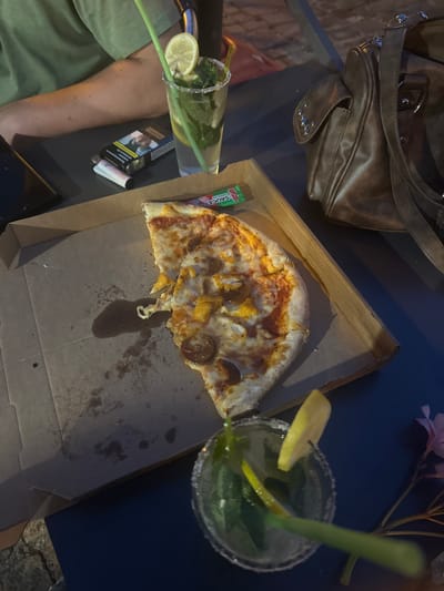 A high-angle, medium shot shows a table in Noyon, France, set for a casual meal outdoors. A partially eaten pizza sits in a cardboard box in the foreground, accompanied by a cocktail with mint and a slice of lemon. A second cocktail, also garnished with mint and lemon, is visible in the background. A brown leather purse rests on the table, and a pack of cigarettes and a lighter are also present. The scene suggests a relaxed atmosphere, perhaps a meal at a cafe or restaurant in the historic city of Noyon.