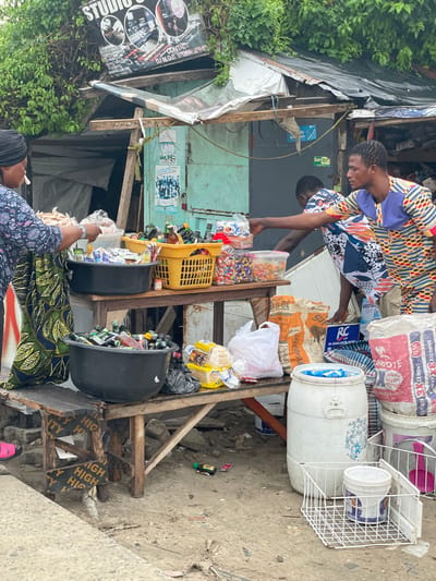 In Ilado, Nigeria, an outdoor market stall displays a variety of goods. A woman, partially visible on the left, is interacting with the stall. She wears a patterned blue and white dress and a black headscarf. In the center of the frame, a man in a vibrant, multicolored patterned shirt is engaged in a transaction with another man, who is partially obscured by the stall. The stall itself is constructed from wood and corrugated metal, with a tarp roof. It is laden with merchandise, including numerous bottles of drinks in a large black basin, a yellow basket filled with bottles, and various packaged goods like candies and snacks. Several sacks, including ones labeled "Supaset" and "Dangote Cement," are visible on the ground. The ground is dirt and concrete, with scattered debris. The environment suggests a bustling, informal marketplace.