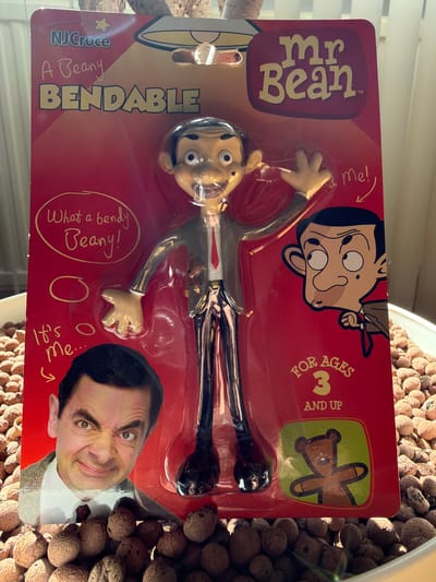 A Mr. Bean bendable toy on red packaging is the primary subject of the image. The packaging includes cartoon images of Mr. Bean and his teddy bear, Teddy. The toy is displayed against a background of small, round, brown clay pebbles. The photograph is taken indoors. There is no information about the location of this image being in the city of Pétange, Luxembourg.