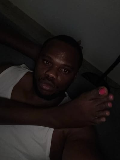 A man is lying on his back, with a foot placed near his head. The man is wearing a white tank top. The foot has pink nail polish on the big toe. The setting appears dark and indoors. The location, Iddo Tudunwada, Nigeria, is not evident from the image itself.