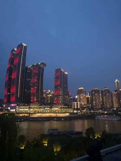 The image depicts a night scene of a modern city skyline with towering buildings illuminated by lights. The perspective is from a slightly elevated viewpoint, looking down at the cityscape and a body of water. Several skyscrapers are prominently featured, some with red illuminated sections. A river or canal runs alongside the buildings, reflecting the city lights. The sky is a deep blue, indicating it is nighttime. The overall mood is serene and awe-inspiring, showcasing the grandeur of urban development at night. There is no visible text in the image. The setting appears to be urban, likely in a developed area with modern architecture. The time of day is clearly night due to the darkness of the sky and the illumination of the buildings.