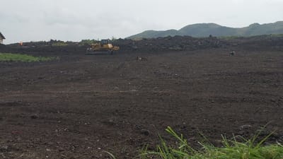 A yellow bulldozer is operating on a large expanse of dark, tilled soil under an overcast sky. Mountains are visible in the background, contrasting with the relatively flat terrain in the foreground. Some grass is visible in the very front and a small structure is to the left.