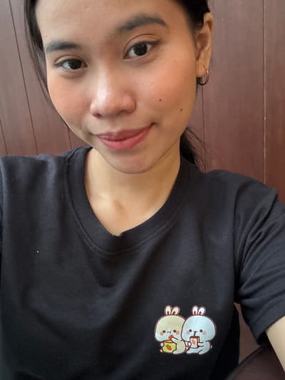 A young woman with dark hair and a black t-shirt with a cute bunny graphic is shown in a close-up selfie. Her face is turned towards the camera, and she is smiling slightly. The background appears to be a wooden wall. The image was taken in South Jakarta, Indonesia.