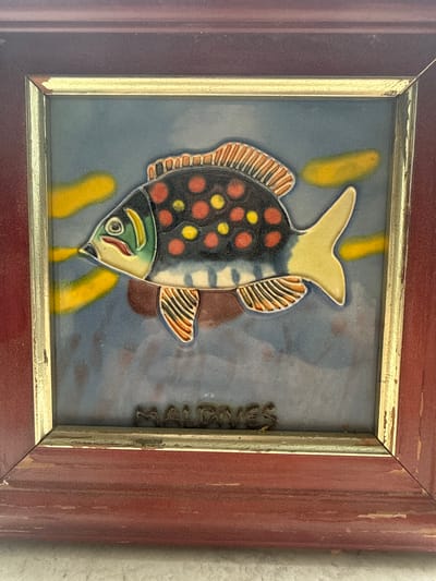 A framed ceramic tile depicting a fish. The fish is colorful, with a black body covered in red and yellow dots, a yellow tail, and orange and yellow fins. The background of the tile is a blue-gray color with yellow streaks. The frame is made of wood and is red with a gold trim. The tile has a metal plaque that appears to say "Maldives". The image shows detail of paint loss and general wear on the frame. There is no evidence to determine this image was taken in Lesnoi Gorodok, Russia.