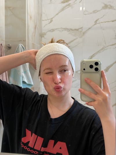 A young individual, appearing female, with reddish-blonde hair secured by a white terrycloth headband, is captured in a mirror selfie. Her face is visibly shiny, consistent with the application of a skincare product. She is winking with her right eye (viewer's left) and puckering her lips in a kissing gesture, while looking towards the right. White wireless in-ear earbuds are present in both ears. Her left hand is raised to touch the headband on the left side of her head, while her right hand holds a light-colored smartphone with a clear case, reflecting the mirror. The smartphone's rear camera array, featuring three large lenses and a smaller sensor, is clearly visible. She is wearing a black t-shirt with a bold red graphic, which includes a chain link pattern, on the chest. A thin silver-colored chain necklace is visible around her neck.
The background, visible in the mirror's reflection, consists of a bathroom interior with light-colored, possibly beige or white, tiled walls featuring a subtle marble-like veining pattern. Two towels, one light pink and one light blue with a white pattern, hang on hooks in the background. The specific location, including city and country, is not discernible from the visual information provided.