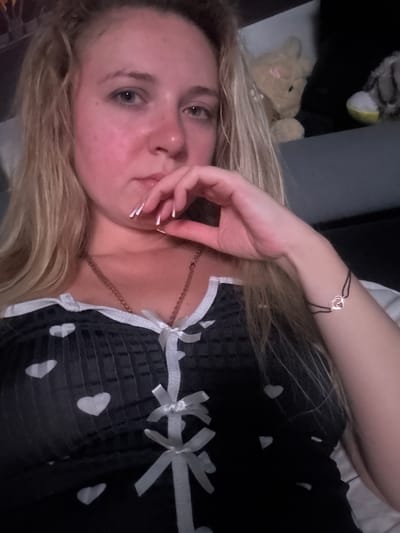 The image shows a close-up, vertically oriented selfie of a young woman with blonde hair. She is lying down, and her face is turned towards the camera, with her hand touching her mouth. She is wearing a black garment adorned with white hearts and white ribbon bows. A delicate bracelet with a heart charm is visible on her wrist. In the background, a stuffed bunny and other indistinct objects are visible, suggesting a casual or personal setting. The lighting appears to be dim, possibly indicating nighttime or an indoor environment. The woman's expression is somewhat pensive or playful. There is no visible text in the image. Given the provided location context of Makiivka, Ukraine, it is likely this image was taken there.